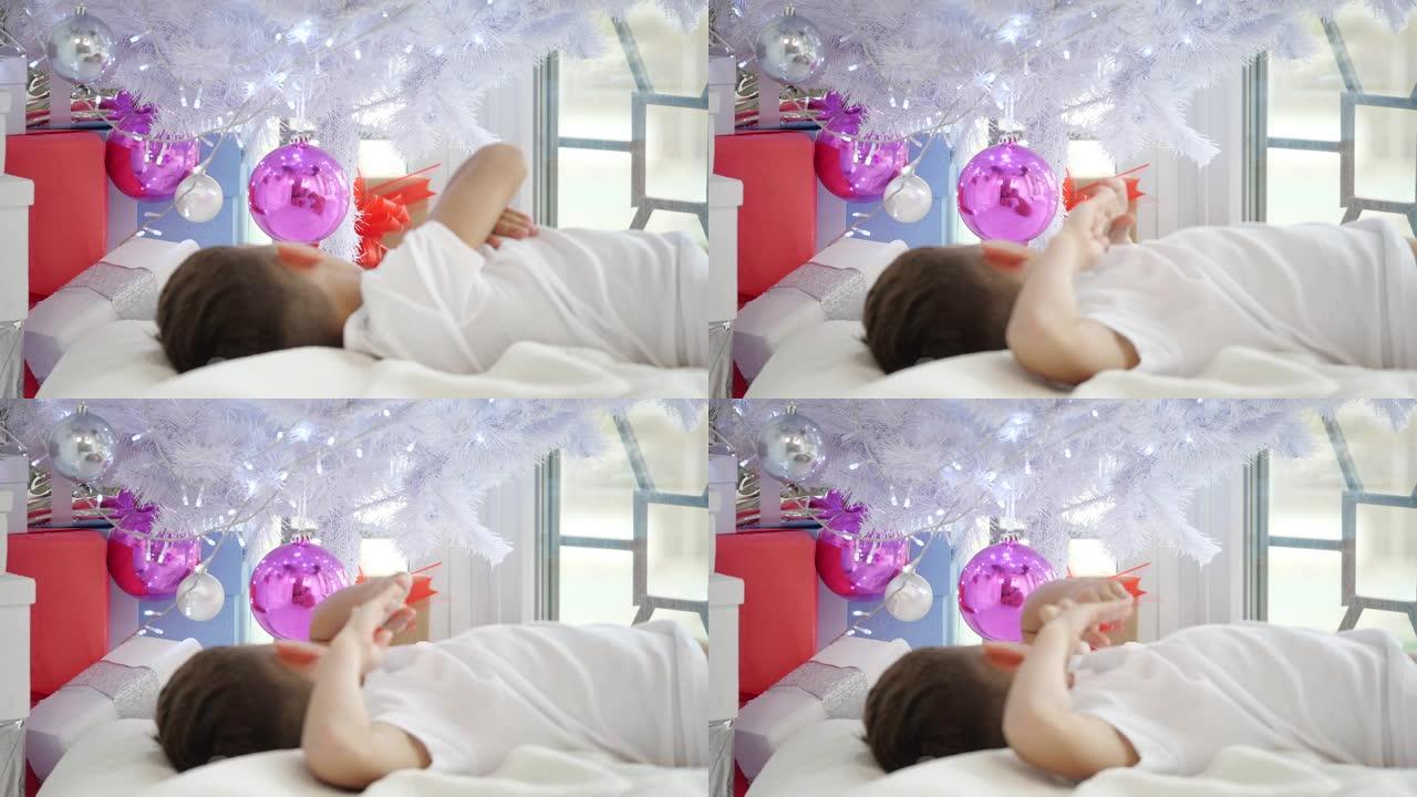 视频的小孩躺在圣诞树下，触摸球，看着他的倒影在粉红色的玻璃球。