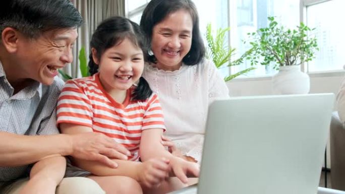 爷爷奶奶教小儿子孙女用笔记本电脑坐在沙发上，幸福的关系结合一代亚洲家庭花时间在网上玩笔记本电脑在家里