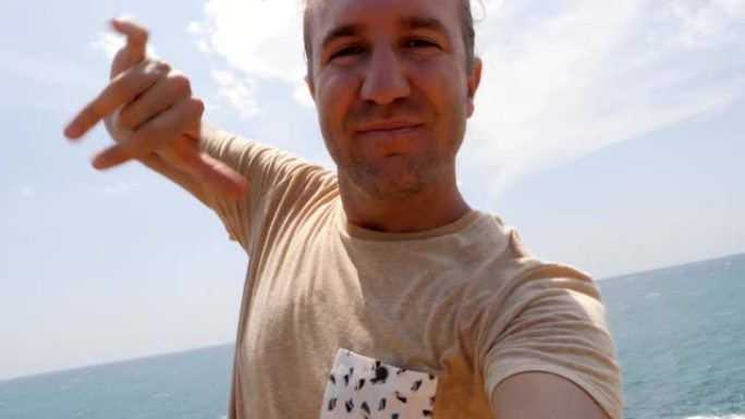 一个有趣的年轻人在悬崖上拍自拍视频，他在海边挥手打招呼，还做了沙卡手势。人们在网上分享度假的概念