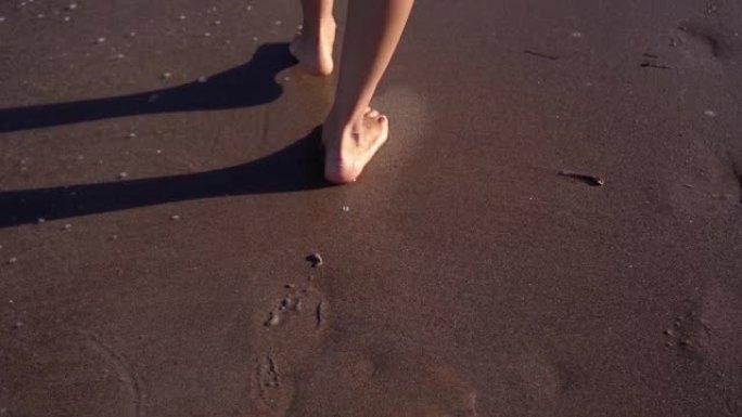 年轻女子的脚沿着海浪边缘附近的湿沙行走