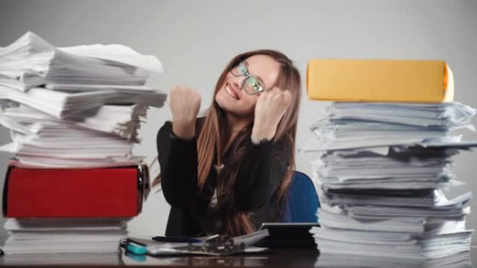 微笑的女办公室经理显示幸福的情绪坐在两堆完成的文件。成功的总经理在镜头前举手示意，心情愉悦。过度工作
