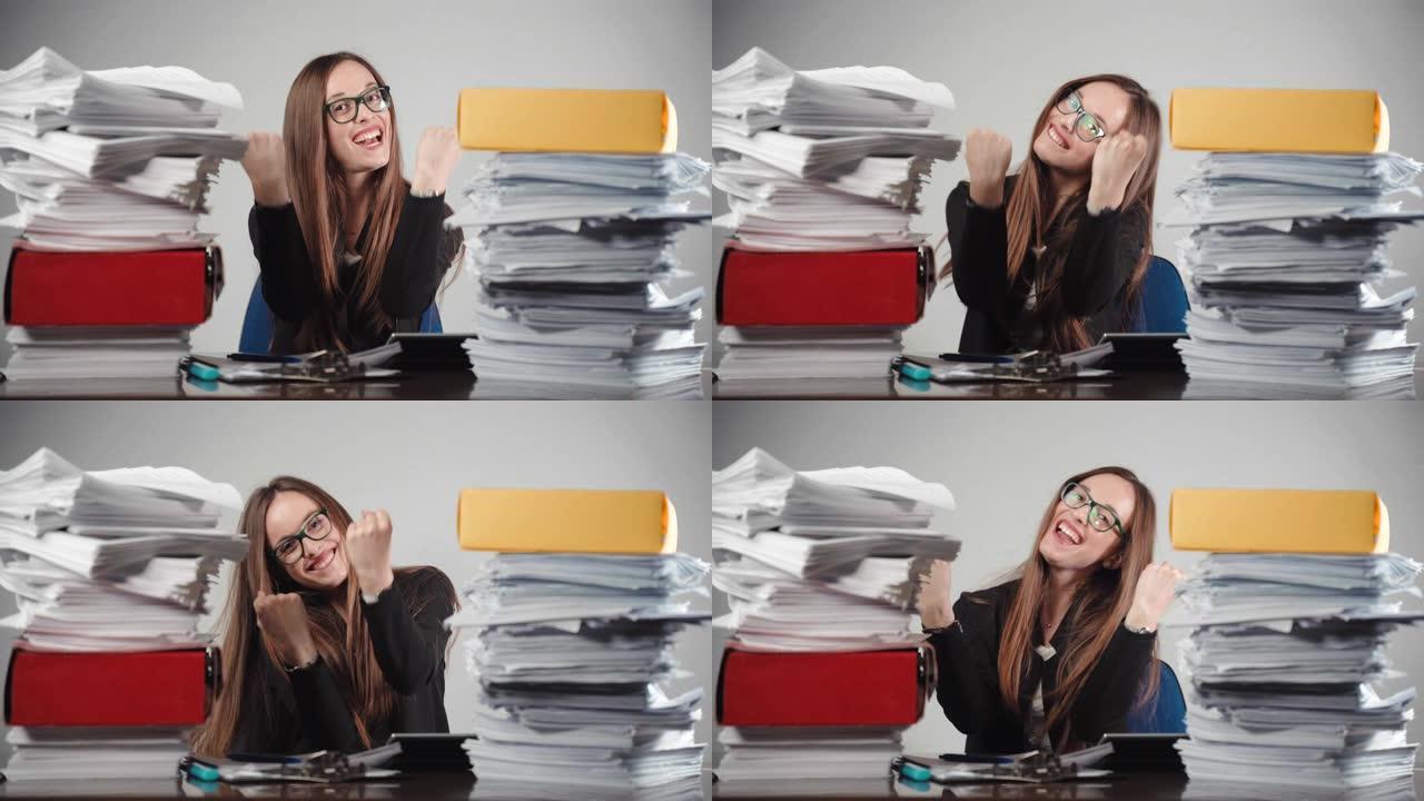 微笑的女办公室经理显示幸福的情绪坐在两堆完成的文件。成功的总经理在镜头前举手示意，心情愉悦。过度工作