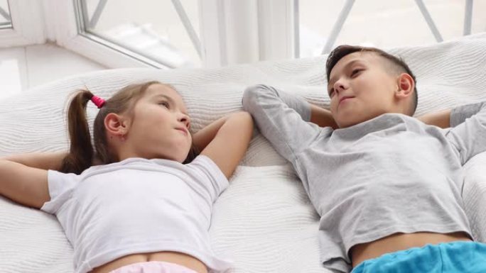 两个美丽的兄弟姐妹一起躺在床上，在休息时微笑和交谈的俯视图。