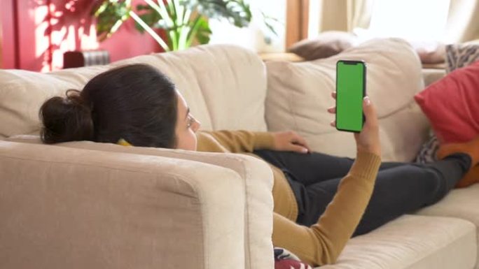年轻的印度妇女躺在沙发上，手里拿着空白的数字电话，手机绿屏，垂直模式下的模拟屏幕