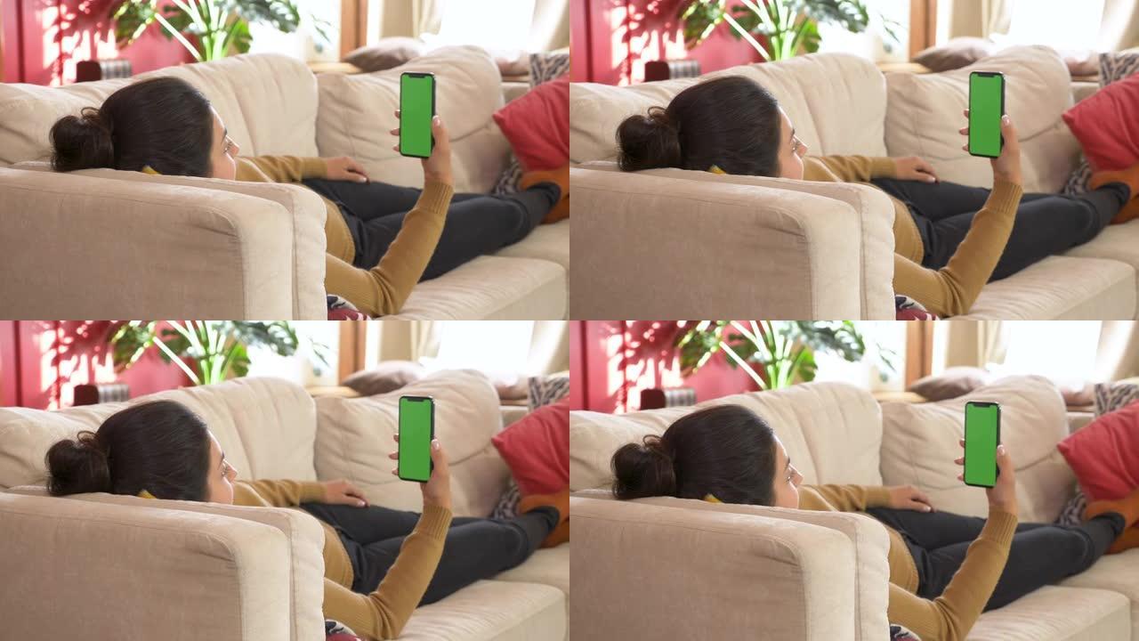 年轻的印度妇女躺在沙发上，手里拿着空白的数字电话，手机绿屏，垂直模式下的模拟屏幕