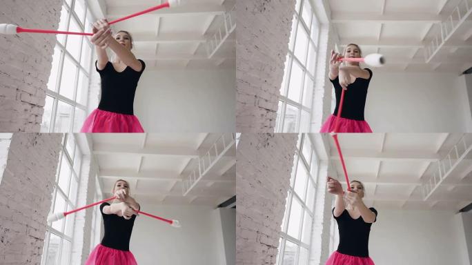 艺术优美的体操运动员手持马鞭子，在体操大厅的白色背景上进行杂技动作。女孩穿着黑色的身体和粉红色的裙子