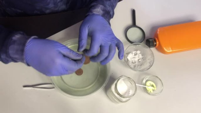 一个戴着橡胶手套的蒙面人从磷酸中拉出铜币。检查并放回。在苏打水和硫酸软膏旁边清洗铜币。