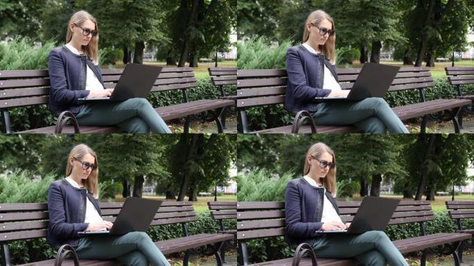 年轻迷人的女商人戴着太阳镜坐在城市公园的长凳上，在笔记本电脑上工作