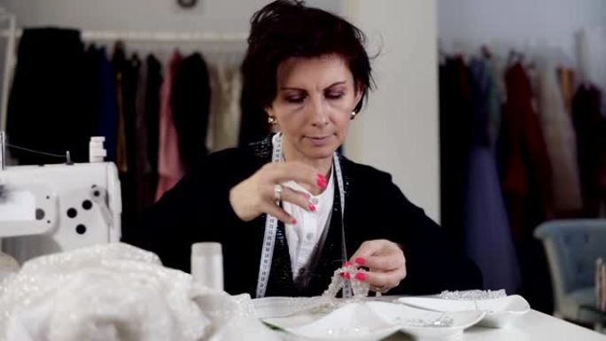 一位女裁缝在婚纱细节上的肖像。拿着针线，用装饰石头装饰独家婚纱。缝制优雅的婚纱。手工制作。裁缝工作室