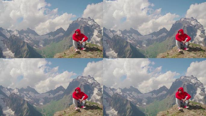 阳光明媚的天气里，戴着墨镜的年轻人坐在山上。穿着红色连帽衫的成年男性，带兜帽，在山区欣赏美丽的景色