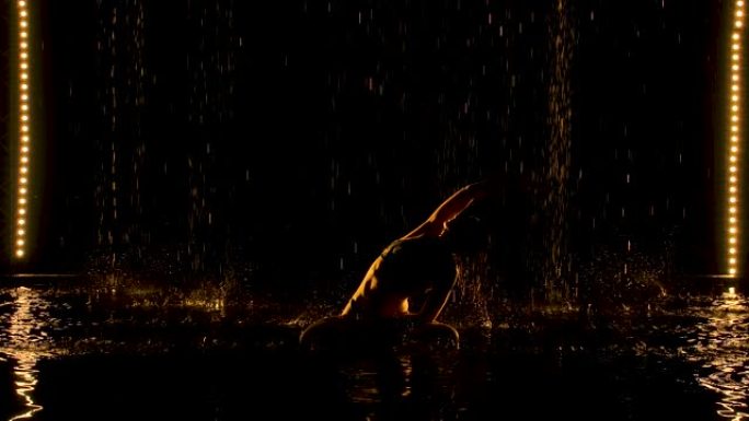 练习瑜伽练习的苗条女人的轮廓。雨滴从天花板上掉下来。黄色的演播室灯光在水面和身体上都是柔和的。慢动作