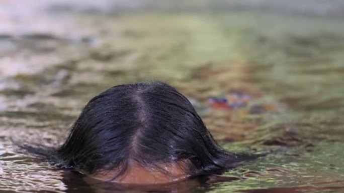 年轻的亚洲妇女在室外游泳池的喷泉喷雾下在水中游泳。