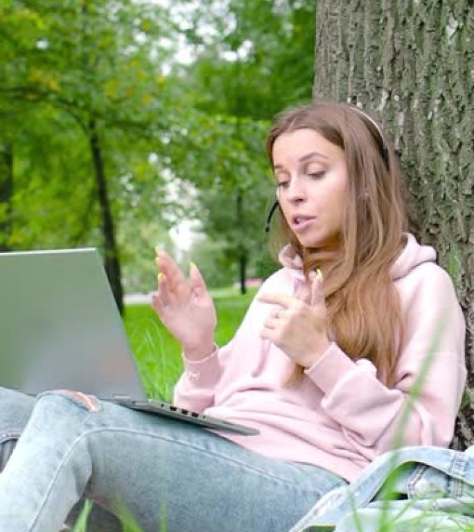 垂直视频-坐在城市公园里的带笔记本电脑和耳机的年轻女子在线对话