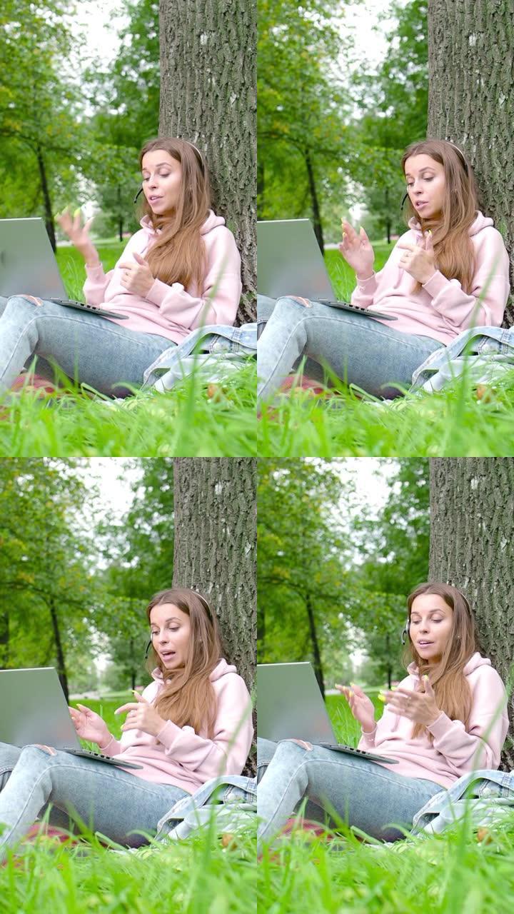 垂直视频-坐在城市公园里的带笔记本电脑和耳机的年轻女子在线对话