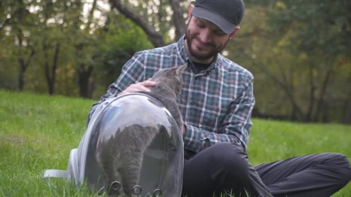 男人使用带舷窗的背包作为宠物，坐在公园的绿草地上玩耍，和灰猫一起玩。设备是透明的呼吸胶囊，有趣的猫从