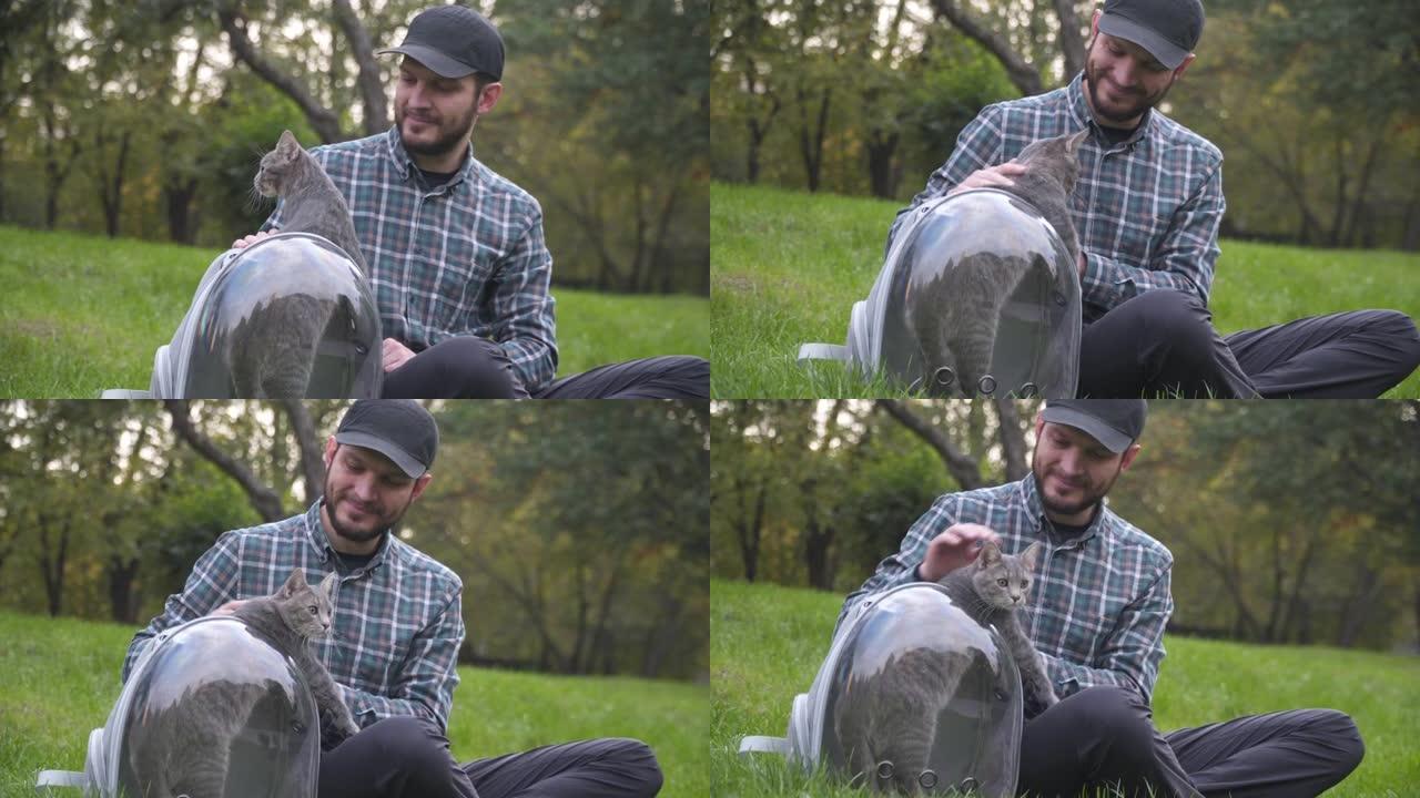 男人使用带舷窗的背包作为宠物，坐在公园的绿草地上玩耍，和灰猫一起玩。设备是透明的呼吸胶囊，有趣的猫从