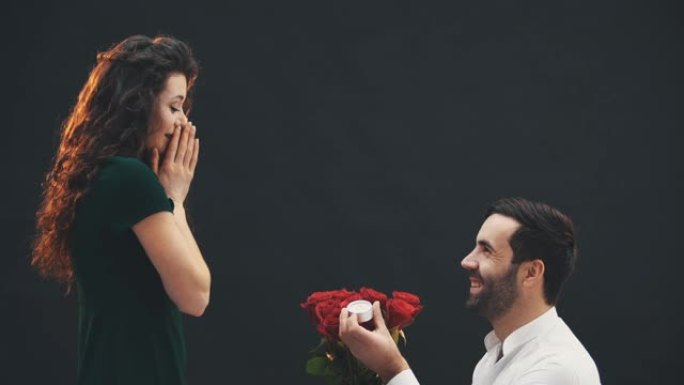 英俊的男人单膝站着，手里拿着戒指和玫瑰，向他的女人求婚。她答应了。
