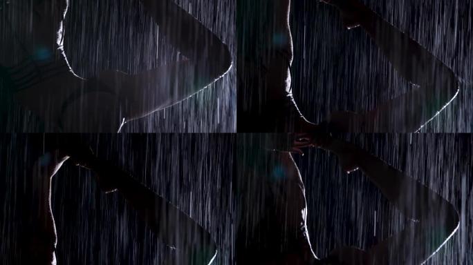 在倾盆大雨中，一名年轻女子表演瑜伽体式。水滴和工作室光线中细长身体的轮廓。黑色背景。特写