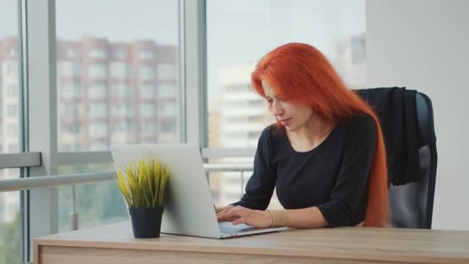 白人外表和红头发的年轻迷人女人在办公室用笔记本电脑工作。女人屏住呼吸，思考和工作。