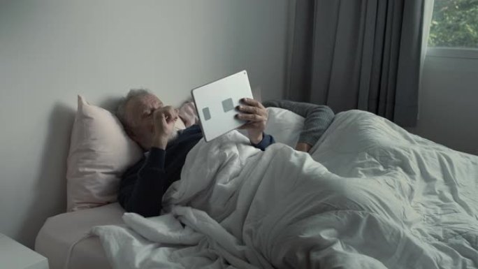 快乐的老年夫妇高加索老人看体育、游戏、电视，在平板电脑上使用，老太太晚上睡在卧室的白色毯子里