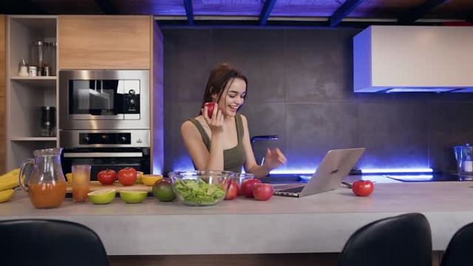 美丽昂扬积极向上的年轻女子在设备齐全的厨房里吃新鲜苹果和使用笔记本电脑