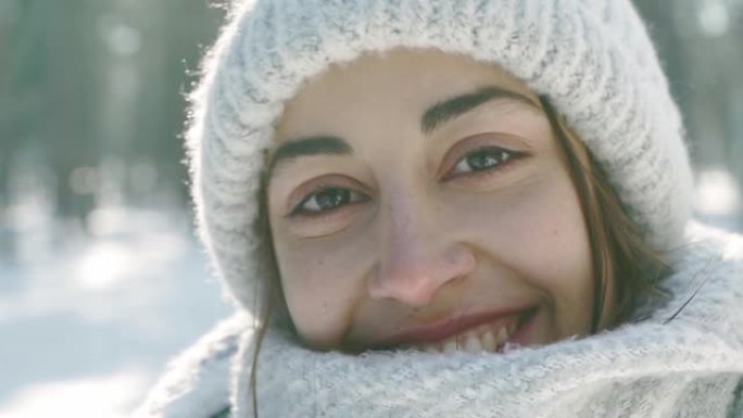 在寒冷的晴天，白雪皑皑的冬季公园里，穿着温暖的羊毛帽和长围巾的美丽大笑的年轻女子的特写肖像。女人看起