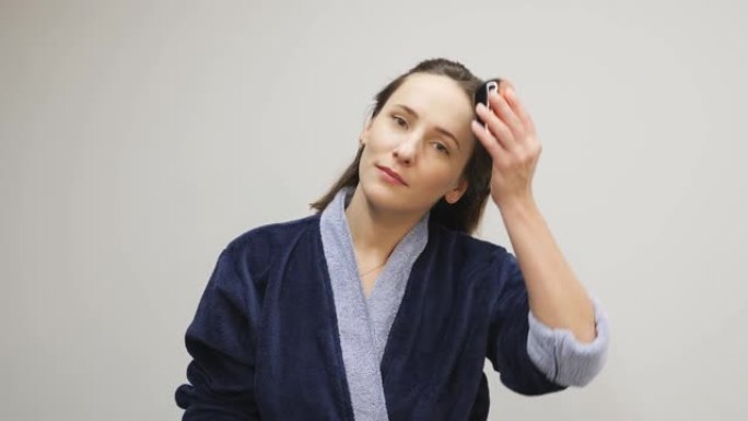 女人在白色背景下用发刷梳理头发。穿着蓝色浴袍的年轻自信女性在睡觉前梳理头发。健康概念