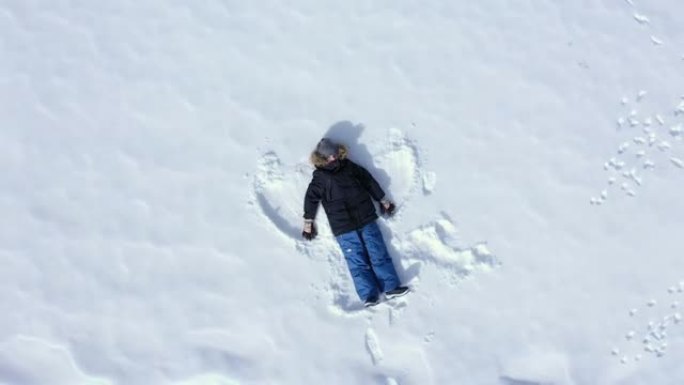 快乐的男孩躺在闪闪发光的雪地上，挥舞着他的胳膊和腿，使雪天使的鸟瞰图滑动。孩子穿着暖和的冬装在雪地里