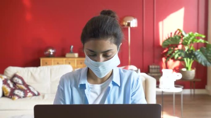 一个戴着口罩的年轻印度妇女的特写镜头，使用笔记本电脑在网上工作，公寓里明亮舒适的房间
