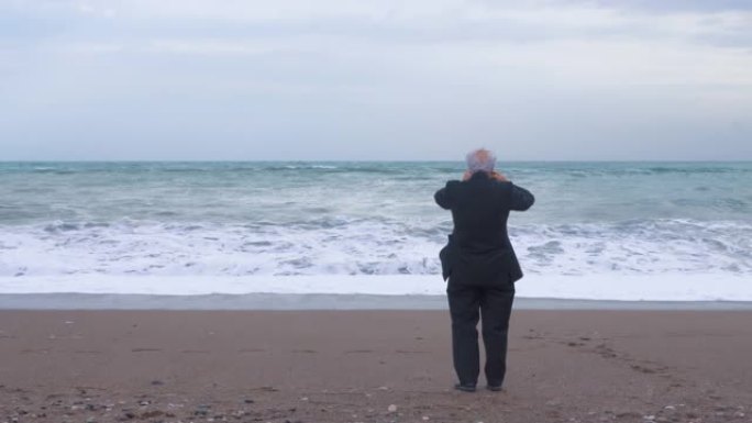 穿着西装的白发老人在恶劣的大风天气中独自站在暴风雨的海岸上。退休人员的抑郁和孤独
