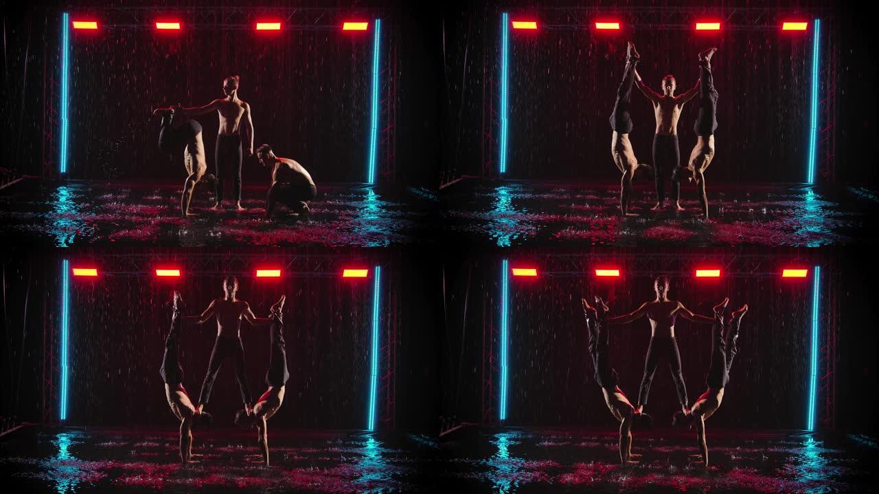 男人在雨中表演杂技特技。三位男杂技演员的专业表演。黑色背景，红色霓虹灯。慢动作