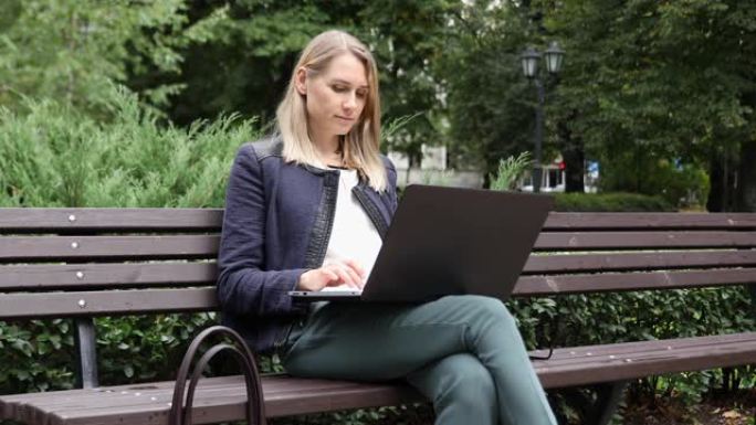 年轻迷人的女人坐在公园的长凳上，使用笔记本电脑