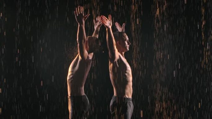 两名体操杂技演员在雨中保持平衡。强壮的运动员在团队合作中表演困难的技巧。在黑色背景上拍摄，慢动作照明