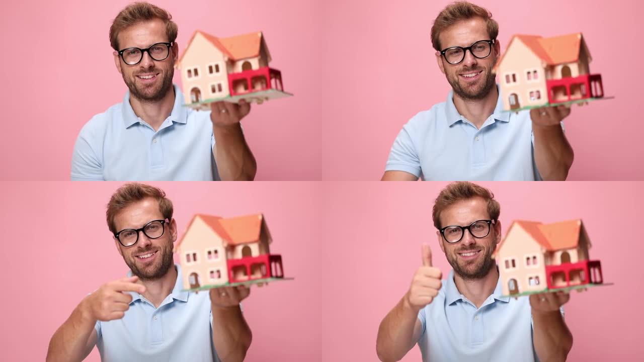 穿着蓝色马球衫的快乐年轻休闲小伙子戴着眼镜，展示房子，指着手指，竖起大拇指，在粉色背景上点头微笑