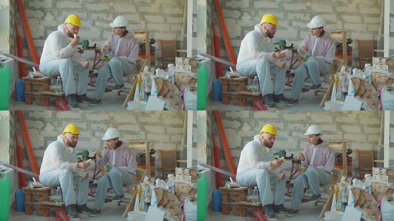 两名画家工人在建筑工地吃午饭。戴着安全帽的男人和女人坐在建筑材料旁，边吃饭边聊天。
