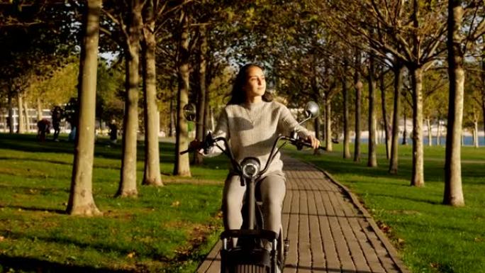 骑着电动滑板车的年轻女孩在阳光明媚的早晨骑着马穿过公共公园