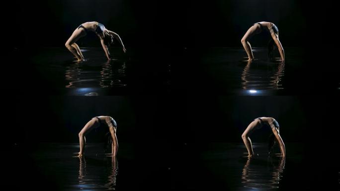 苗条的女人穿着湿运动内衣在水面上练习桥式锻炼。性感的身体在工作室的灯光下闪闪发光。黑暗工作室。黑色背