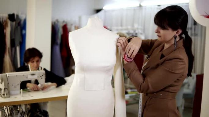 黑发女人把布袖钉在模特身上，另一个女人在现代工作室的背景下在缝纫机上工作。女士们专注于她们的工作