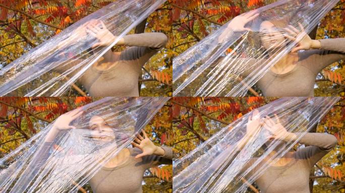 金发短发的年轻女子正透过塑料袋，聚乙烯包裹站在户外，在阳光明媚的秋天，靠近黄色和红色的叶子的树。