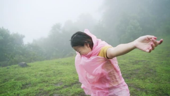 穿着粉红色雨衣的快乐年轻亚洲女性旅行者的慢动作享受和乐趣，在雨中与美丽的大自然一起旅行。微笑的漂亮女