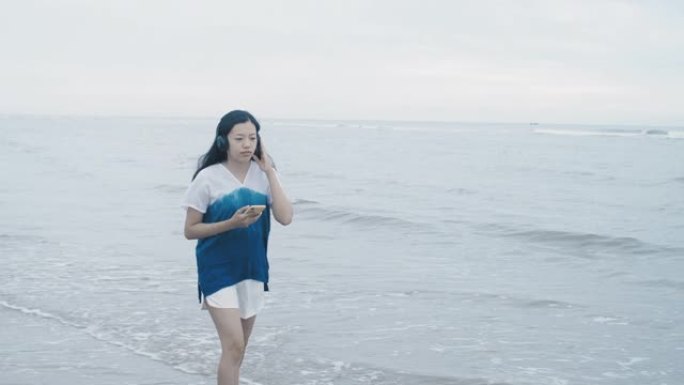 亚洲美女周末度假时，在海边的海滩上使用带耳机的智能手机听音乐。快乐的单人旅行概念。