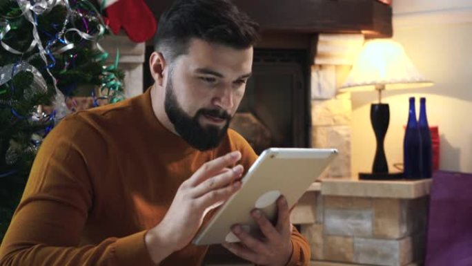 集中高加索人的肖像，黑胡子坐在圣诞树前，使用平板电脑。严肃的年轻人在除夕夜网上搜索礼物。假期，惊喜，