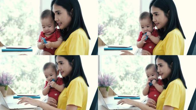 美丽的商业亚洲妈妈正在使用笔记本电脑，微笑着与她可爱的宝宝在一起。休产假的年轻母亲试图带着蹒跚学步的
