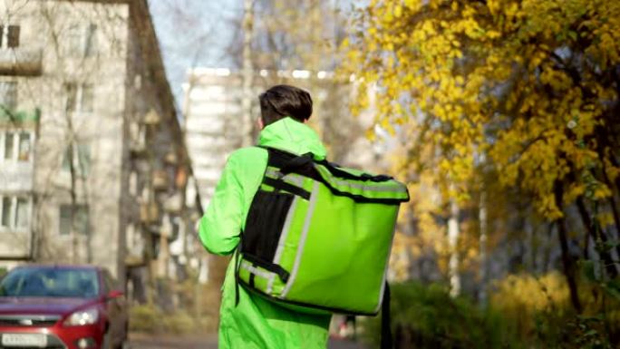 使用智能手机上的移动导航应用程序在秋季街上搜索方向的绿色制服和保温袋年轻食品快递员的后视镜头