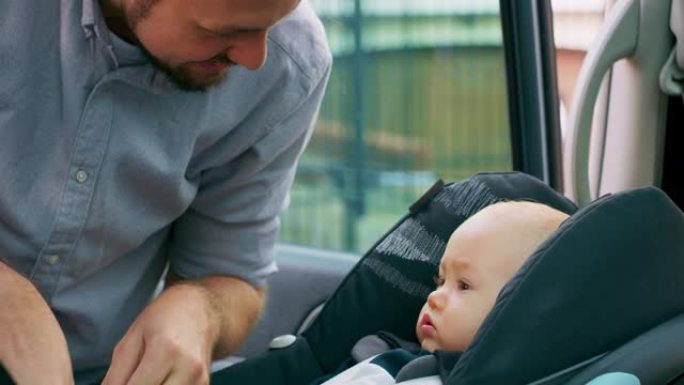 车内摄像头。特写男婴坐在车内的婴儿汽车座椅上。年轻的大胡子父亲检查婴儿系好的安全带，微笑着与他交谈，