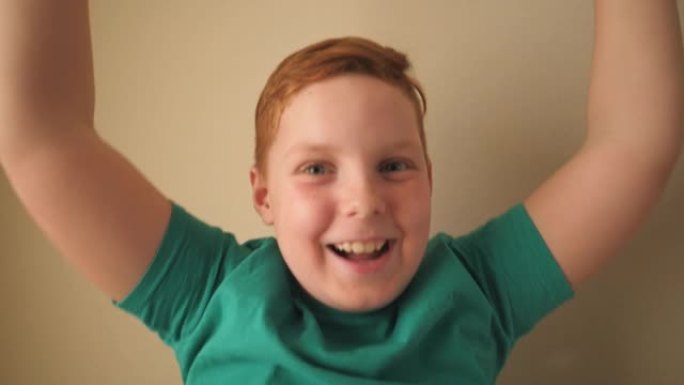 快乐的红发小男孩带着雀斑看着镜头，举手欢喜室内成就。男性姜孩子的肖像，脸上充满积极的情绪。慢动作