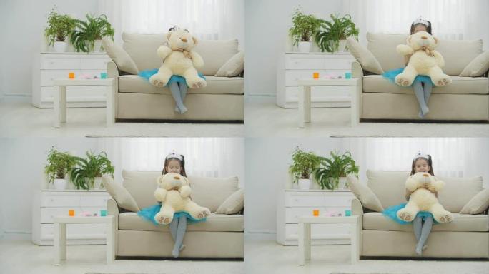 穿着皇冠和礼服的小公主坐在沙发上，躲在泰迪熊后面，挥舞着拍掌。