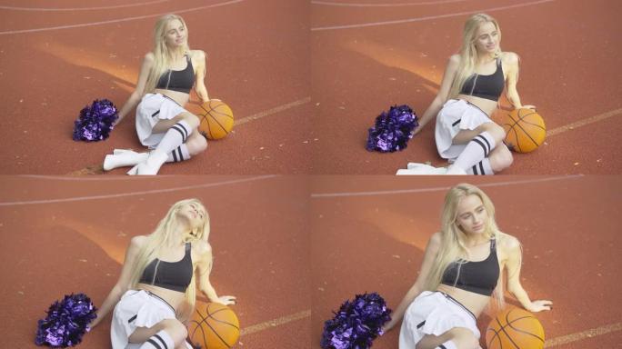 快乐的年轻苗条的啦啦队长的肖像坐在户外球场上，拿着篮子球和绒球。夏日，微笑的金发白人妇女在阳光下在篮