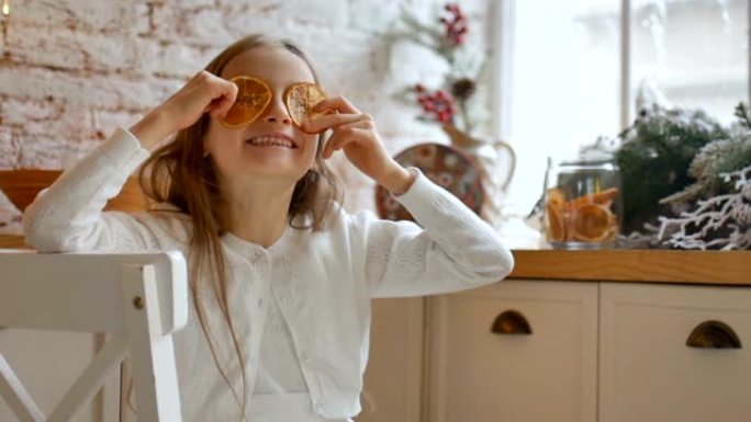 微笑的金发小女孩在家里做鬼脸，用干橙片遮住眼睛，背景是阁楼风格和圣诞装饰，圣诞快乐的概念