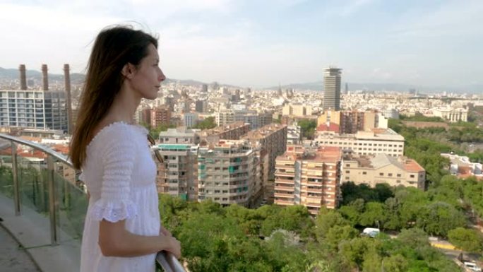 美丽的女人站在蒙特里奇山顶上，看着巴塞罗那的城市景观。西班牙。穿着白色连衣裙的女孩享受着她的暑假。旅